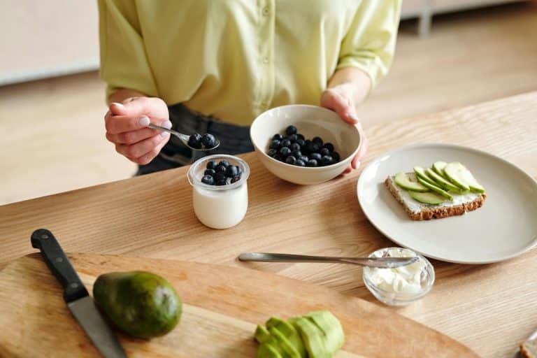 Woman preparing a balanced PCOS snack with Greek yogurt, blueberries, and avocado toast on whole grain bread.