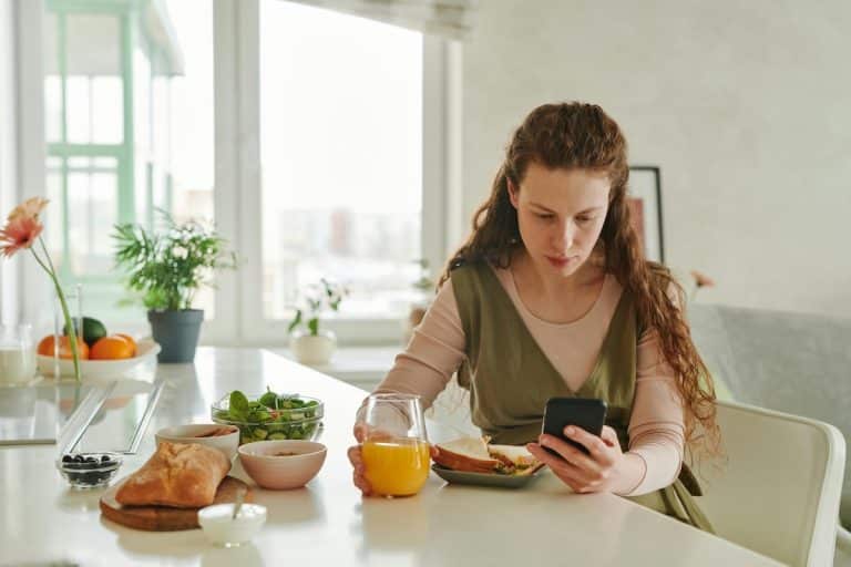 A woman with a serious expression sits alone at a bright kitchen table with a variety of foods, including salad, bread, juice, and yogurt, while looking at her phone. Captures the emotional complexity of eating with PCOS and the hidden struggles of disordered eating.