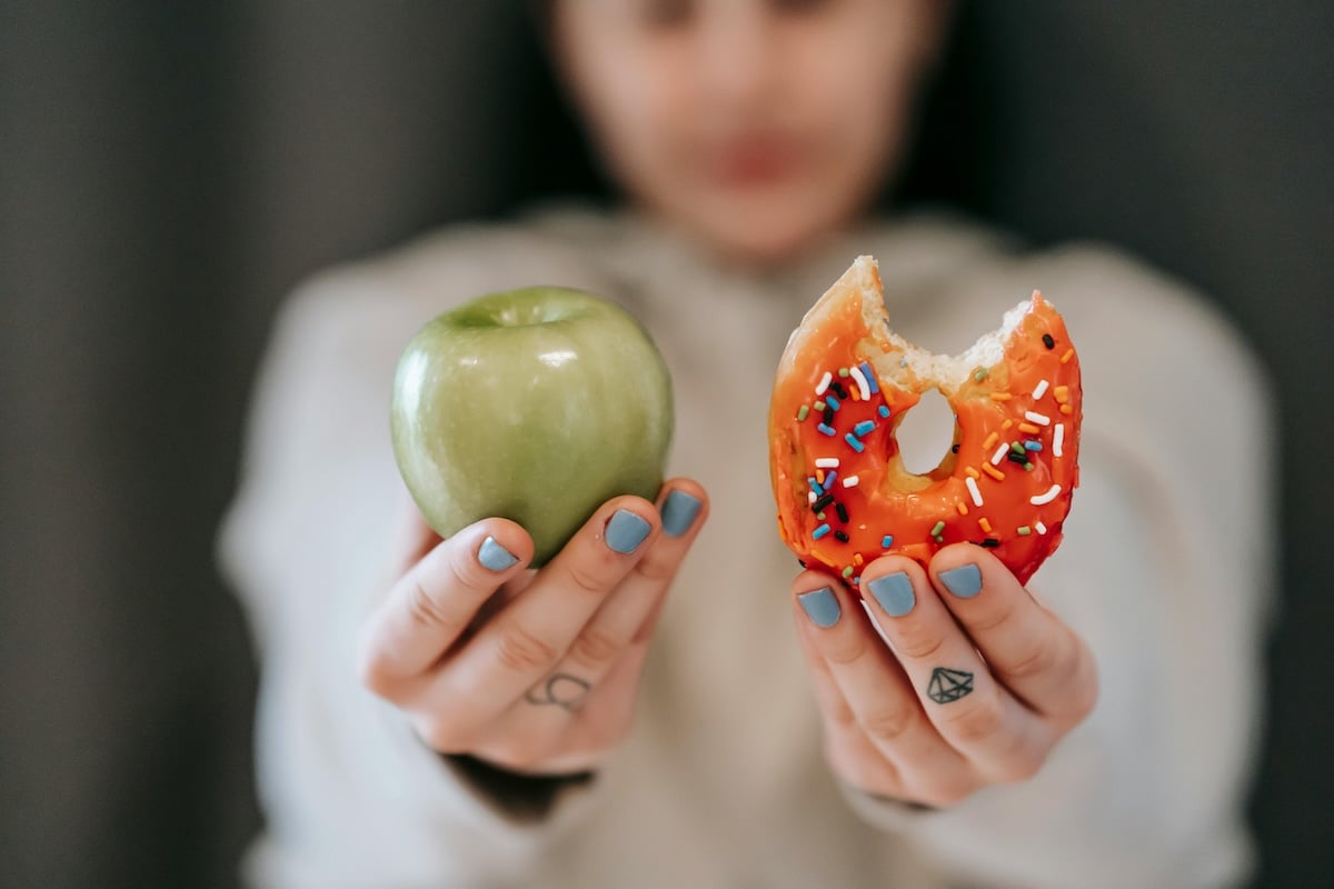 A woman holds an apple and a donut, representing the mental tug-of-war between food restriction and indulgence common in disordered eating with PCOS.