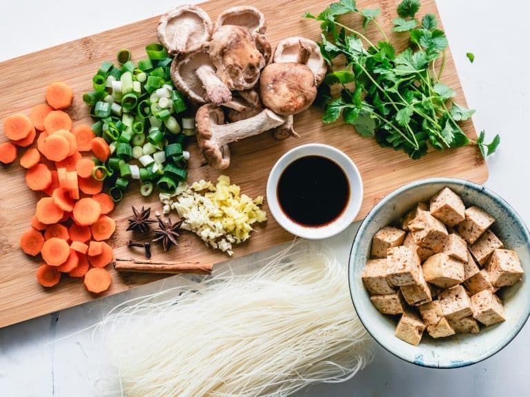 Fresh ingredients for a vegetarian PCOS diet soup, including carrots, shiitake mushrooms, scallions, cilantro, garlic, ginger, tofu cubes, rice noodles, and soy sauce on a wooden board.