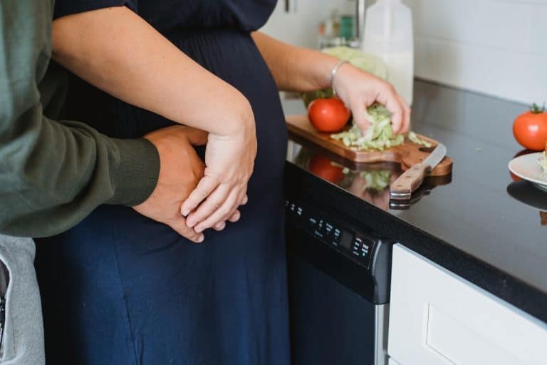 Woman preparing a healthy meal at home while her partner embraces her and holds her pregnant belly—symbolizing the power of fertility foods for PCOS and nourishing the journey to parenthood.